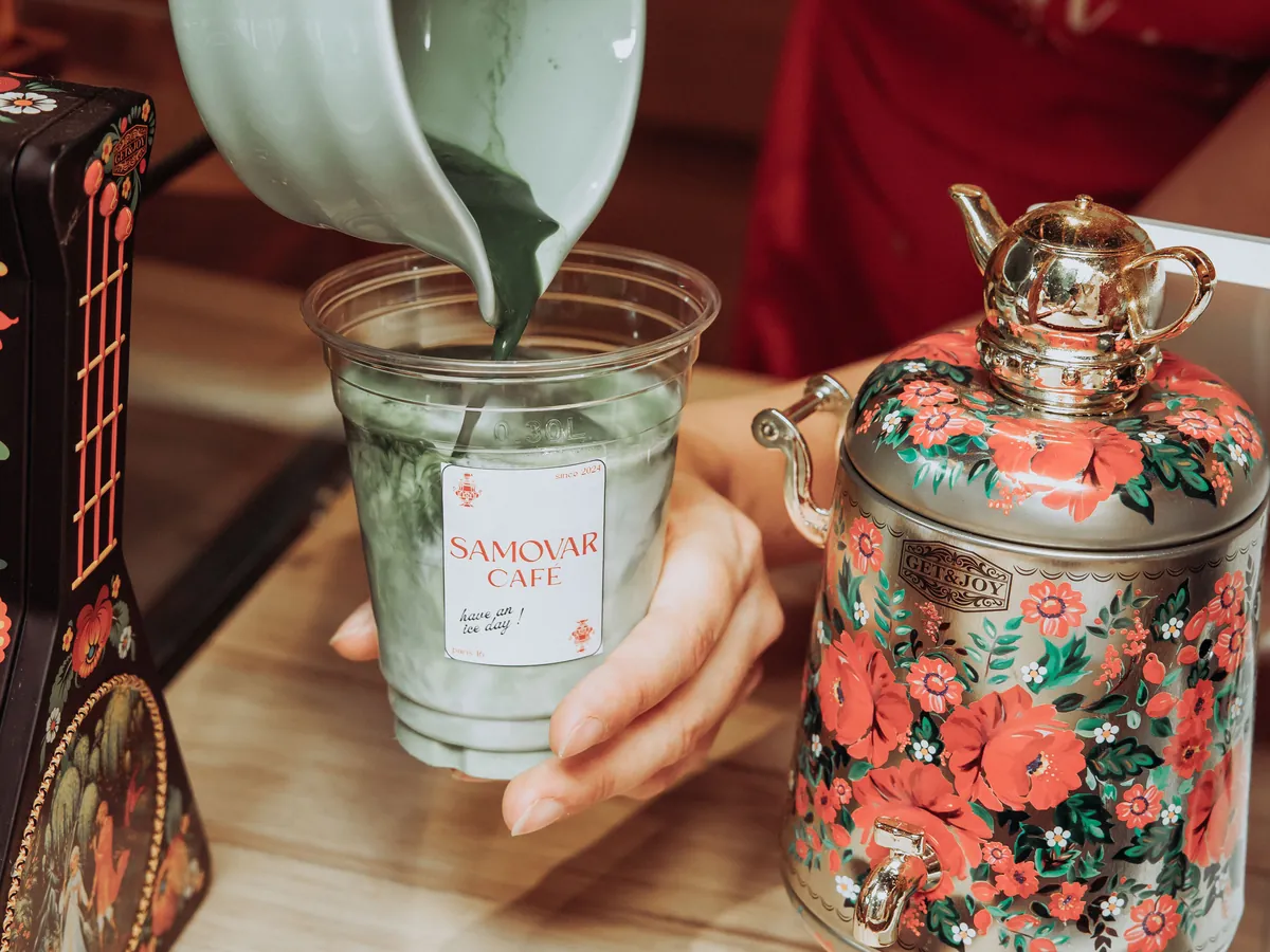 Du matcha versé dans une boisson glacée du Samovar Café, à côté d'un samovar peint de fleurs.
