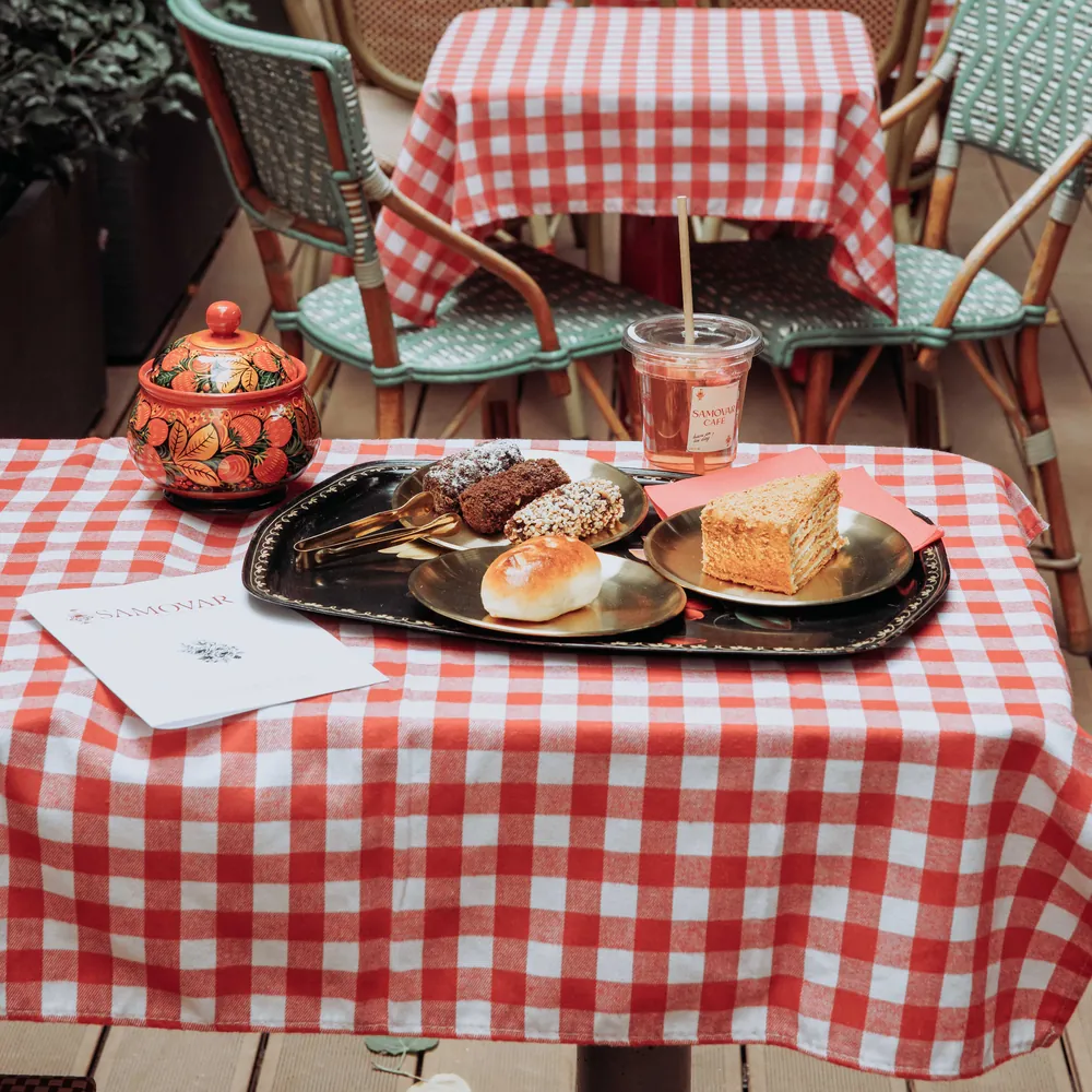 Une table vichy sur la terrasse : medovik, pirojok, gourmandises, un sucrier peint et une boisson glacée.